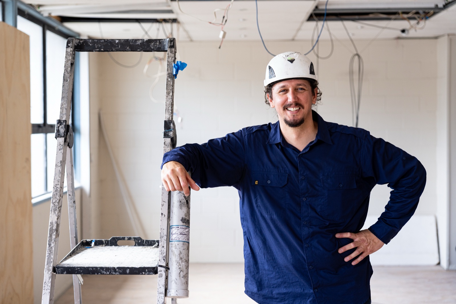 Damyn Blindell of Halt Fire on site in a hard hat, standing beside a ladder in a building under fit-out